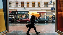Mies mustassa takissa ja keltaisella sateenvarjolla kävelee sadepäivänä jalkakäytävällä