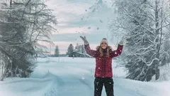 Nainen iloitsee talvisessa maisemassa lumisateessa, lumipeitteiset puut taustalla.