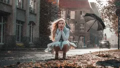 Tyttö istuu kadulla syksyn lehdillä, tuuli lennättää sateenvarjoa taustalla kaupungissa.